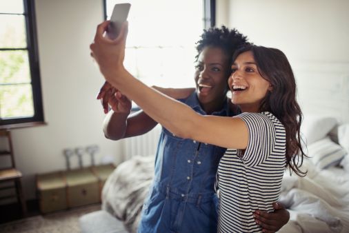 Smiling lesbian couple hugging, taking selfie with camera phone in bedroom