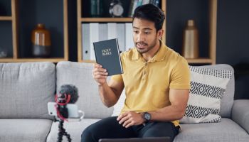 Streaming, bible and man talking on phone and microphone online for live podcast. Asian male on home sofa with Christian religion book as blog content creator or influencer teaching or studying