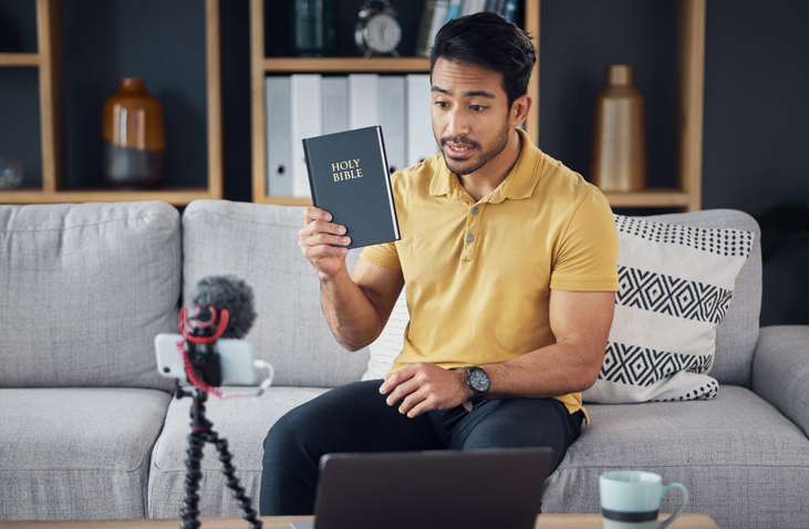 Streaming, bible and man talking on phone and microphone online for live podcast. Asian male on home sofa with Christian religion book as blog content creator or influencer teaching or studying