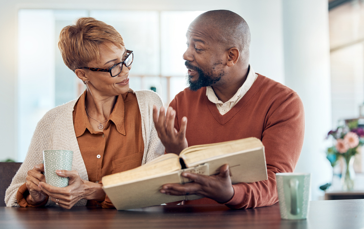 Couple reading bible, home faith and talking about religion with coffee for learning, study and education. Black man, holy book in hand and spiritual conversation about christianity, God and worship