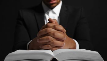 African American man with Bible praying to God at table on black background, closeup