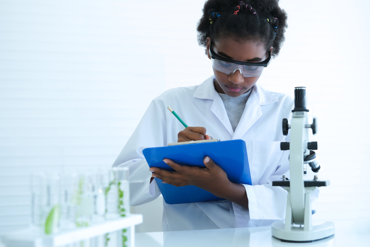 Black curly-haired child scientist holding clipboard to record experiment results at science laboratory.