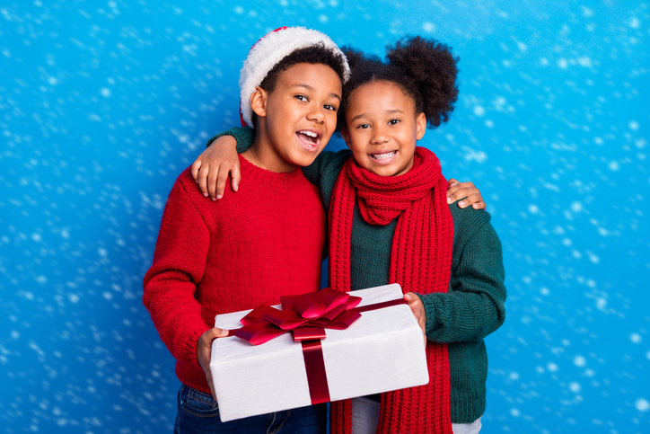 Photo of charming sweet dark skin kids dressed sweaters headwear scarf embracing holding gift smiling isolated blue color background